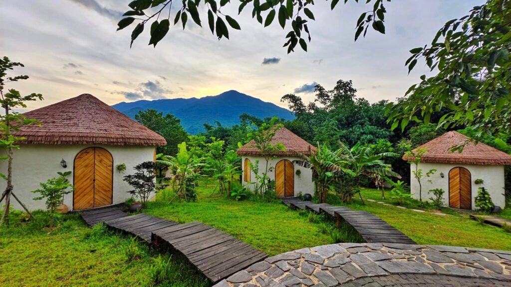 Vì Sao 90% Resort Chọn Rơm Nhân Tạo Làm Mái? Dự Án V Village Đà Nẵng Là Ví Dụ Điển Hình 3 Văn bản thay thế: Lối đi gỗ dẫn đến ngôi nhà mái lá giữa rừng cây xanh mướt