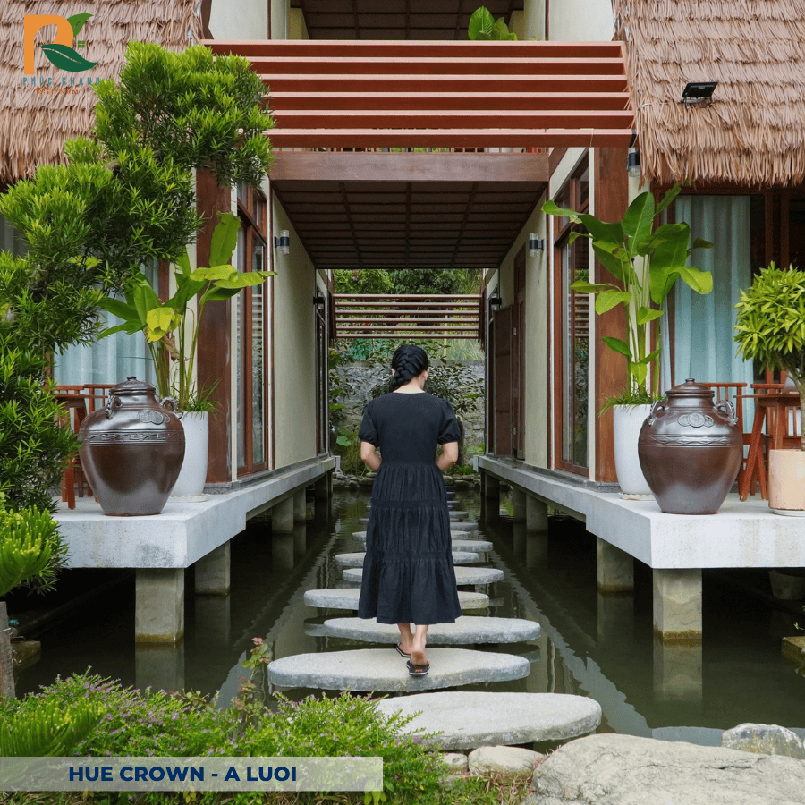 Rơm Nhân Tạo tại Hue Crown – A Lưới: Vẻ Đẹp Truyền Thống Trong Không Gian Nghỉ Dưỡng Xanh 2025 9 Du khách chụp ảnh bên khung nhà gỗ lợp rơm nhân tạo, nền trời chiều tạo hiệu ứng ánh vàng ấm áp – điểm nhấn du lịch sinh thái tại Huế.